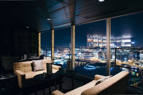 A stylish lounge with modern furniture and a panoramic view of a city skyline at night, illuminated by lights.