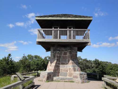 A stone observation tower with a wooden roof, surrounded by greenery and a clear blue sky.