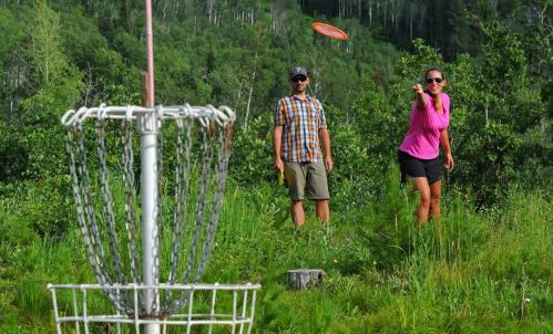 A woman throws a frisbee towards a disc golf basket while a man watches in a green, wooded area.