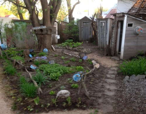 A quaint garden with a winding path, surrounded by rustic sheds and greenery, featuring decorative stones and plants.