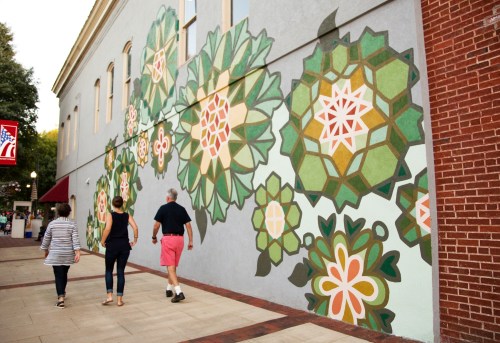 Three people walk past a colorful mural of large flowers on a building wall in an urban setting.