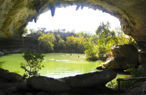 A scenic view of a cave opening overlooking a green pond surrounded by lush trees and people enjoying the water.