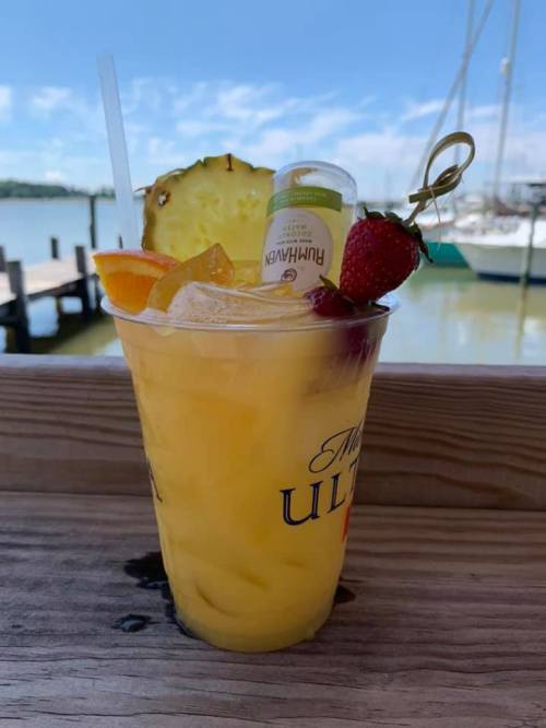 A vibrant tropical cocktail with pineapple, strawberry, and an orange slice, served in a clear cup by the water.