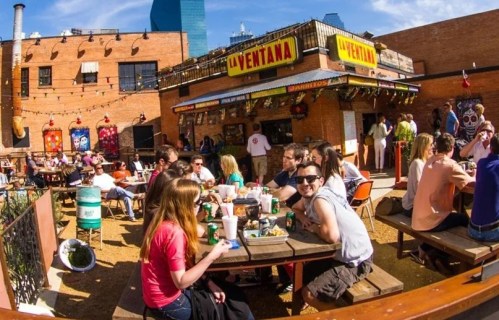 A lively outdoor restaurant scene with people dining at picnic tables, colorful decor, and a sunny atmosphere.