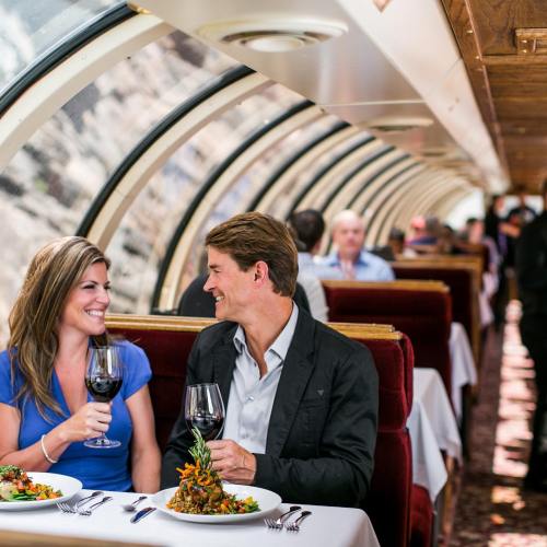 A couple enjoys a meal with wine on a scenic train, surrounded by other diners in a cozy, elegant setting.