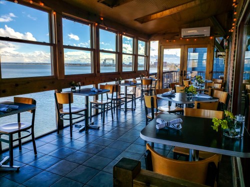 Bright restaurant interior with wooden decor, tables set for dining, and large windows overlooking a scenic waterfront view.