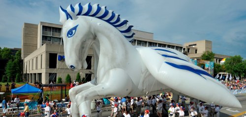 A large white and blue inflatable horse floats above a parade, surrounded by people in white shirts and red hats.