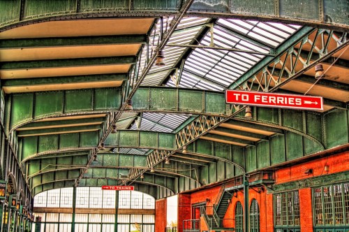 Interior of a train station with green metal beams and signs directing to ferries and trains. Natural light filters in.