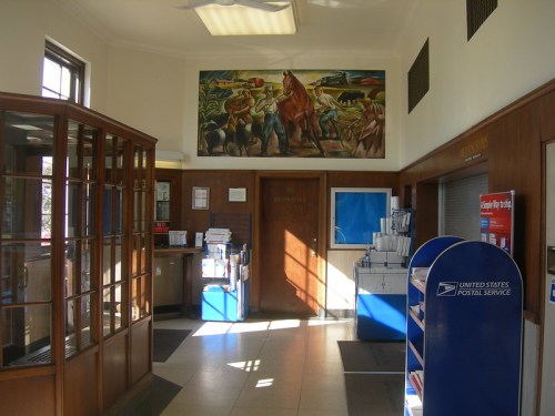 Interior of a post office featuring a mural, wooden counters, and postal service equipment. Bright sunlight streams in.