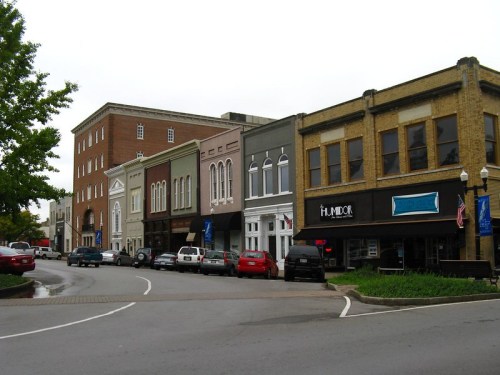 A street view of a small town with historic buildings, parked cars, and trees lining the sidewalk on a cloudy day.