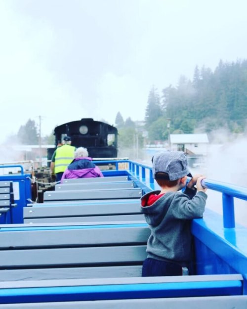 A child in a conductor's hat watches a steam train from a blue boat, with mist and trees in the background.