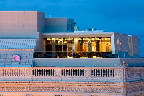 Modern rooftop terrace with glass walls, overlooking a cityscape at dusk, illuminated by warm lights.