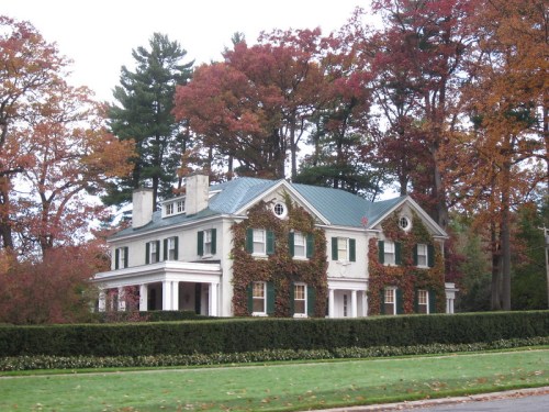 A large, elegant house with green shutters, surrounded by colorful autumn trees and a neatly trimmed hedge.