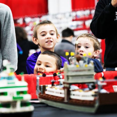 Three young boys smile and watch a display of model ships at a LEGO event, surrounded by colorful decorations.