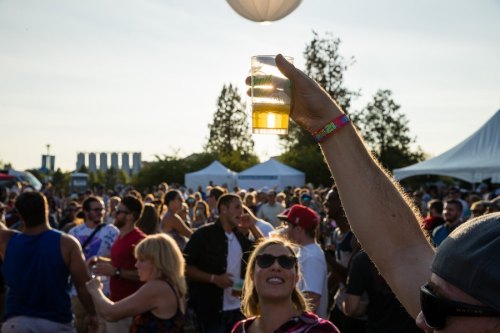 A crowd at an outdoor festival, with a hand raised holding a cup of beer against a sunset backdrop.