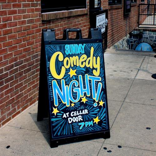 A chalkboard sign advertising "Sunday Comedy Night" at Cellar Door, starting at 7 PM. Bright colors and playful design.