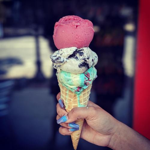 A hand holding a tall ice cream cone with three scoops: pink, cookies and cream, and mint chocolate chip.