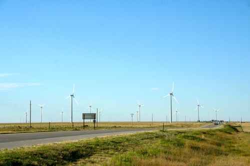 A wide-open landscape with a road, featuring multiple wind turbines against a clear blue sky.
