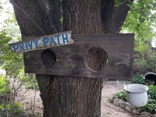 A wooden sign reading "Privy Path" hangs on a tree, with two circular cutouts and a white bucket below.