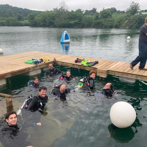 A group of divers in wetsuits is in a lake, with a wooden dock and a shark fin buoy in the background.