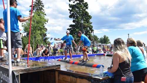 Two participants balance on a log in a water competition, while a crowd watches and cheers from the sidelines.