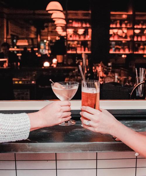 Two hands clink glasses in a dimly lit bar, one holding a martini and the other a tall drink.