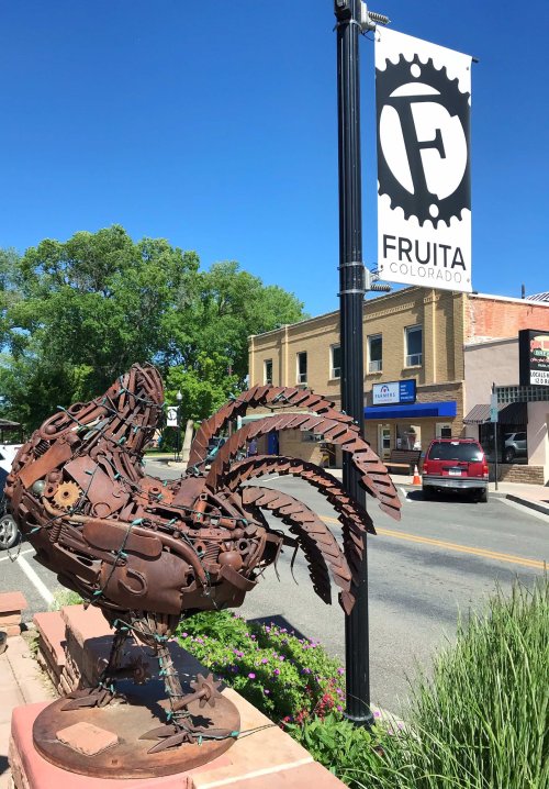 A metal rooster sculpture stands near a street sign for Fruita, Colorado, with shops visible in the background.