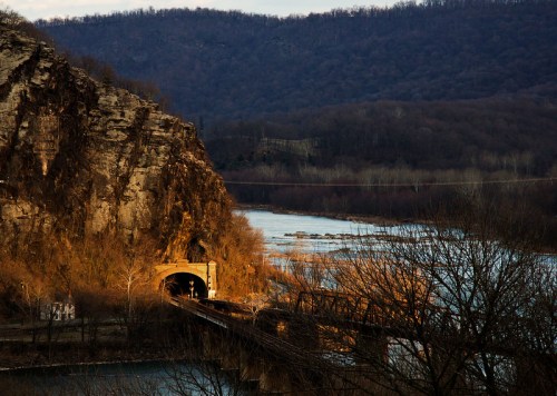A scenic view of a river and rocky cliffs, featuring a tunnel and a bridge under soft evening light.