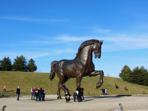 A large bronze horse statue stands tall, surrounded by visitors against a clear blue sky and grassy backdrop.