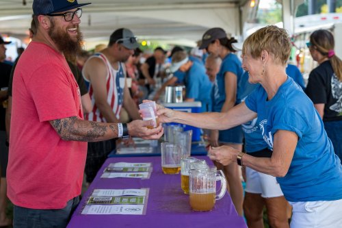 A smiling woman hands a cup of beer to a man at a festival, with others enjoying drinks in the background.
