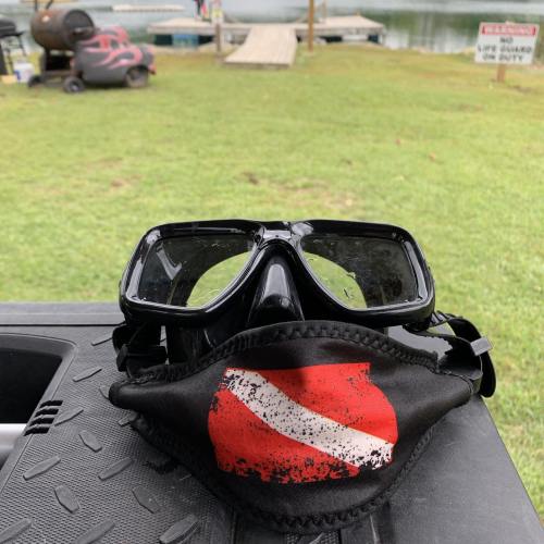 A pair of black diving goggles with a red and white dive flag resting on a table by a lake.