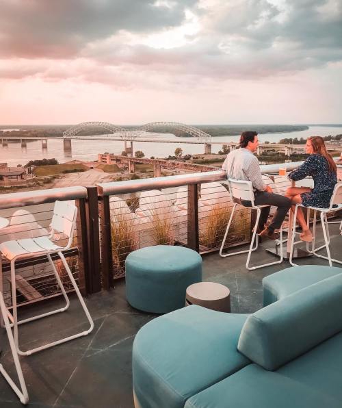 A couple sits on a rooftop bar, enjoying drinks with a view of a river and bridge at sunset.