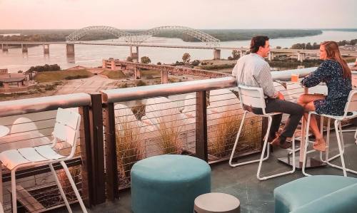 A couple sits at a rooftop bar, enjoying drinks with a view of a river and bridge at sunset.