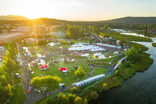 Aerial view of a lively outdoor festival with tents, crowds, and a river, set against a sunset backdrop.