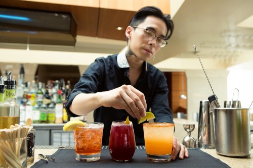 A bartender garnishes three colorful cocktails with lemon slices at a stylish bar.