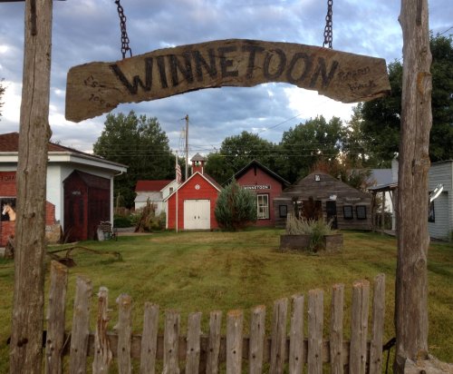 A wooden sign reading "WINNETOON" hangs over a grassy area with buildings and trees in the background under a cloudy sky.