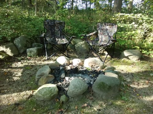 Two camouflage folding chairs sit beside a stone fire pit in a wooded area, surrounded by greenery.