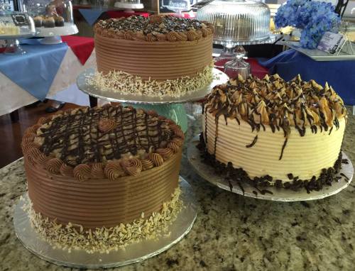 Three beautifully decorated cakes on a countertop: one chocolate with nuts, one with chocolate drizzle, and a vanilla cake with chocolate shavings.