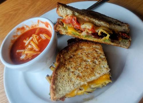 A grilled sandwich with melted cheese and vegetables, served with a small bowl of tomato soup and pasta.