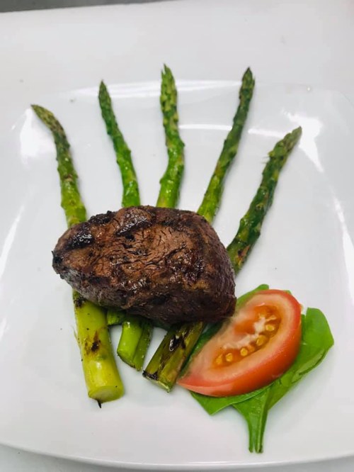 A grilled steak served on a plate with asparagus and a slice of tomato on a bed of leafy greens.