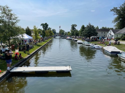 A scenic view of a canal lined with boats, trees, and people enjoying a sunny day at a community event.