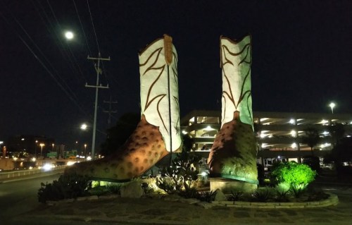 Giant illuminated cowboy boots sculpture at night, surrounded by greenery and a parking garage in the background.