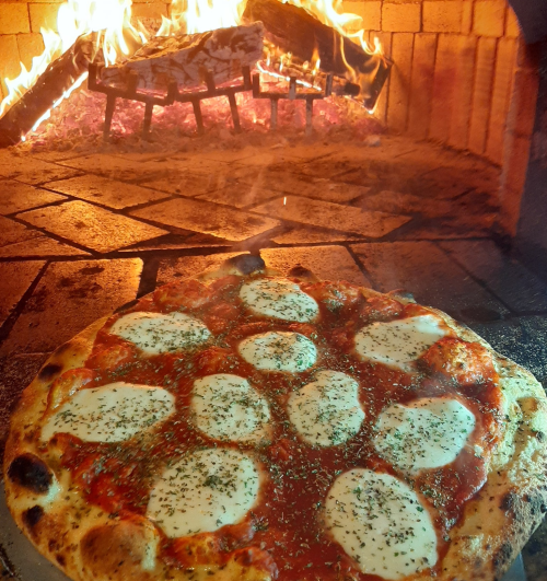 A freshly baked pizza with mozzarella and herbs, placed in a wood-fired oven with flames in the background.
