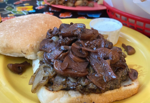 A close-up of a sandwich with a beef patty topped with sautéed mushrooms and onions, served on a yellow plate.