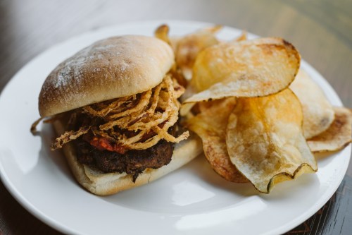A burger with crispy onion strings on a bun, served with a side of potato chips on a white plate.