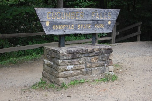 Sign for Cucumber Falls at Ohiopyle State Park, mounted on a stone base, surrounded by greenery.