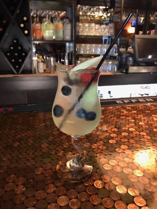 A refreshing cocktail with blueberries and a cherry, served in a decorative glass on a penny-covered bar.