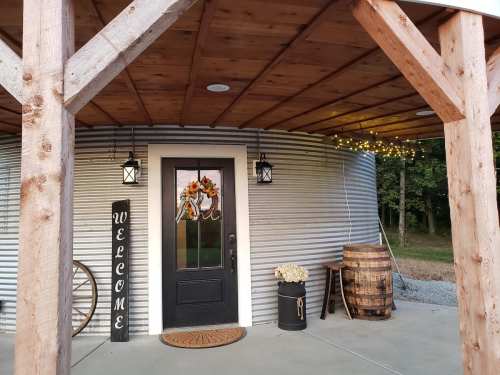 A welcoming entrance with a black door, decorative wreath, lanterns, and rustic decor under a wooden porch.