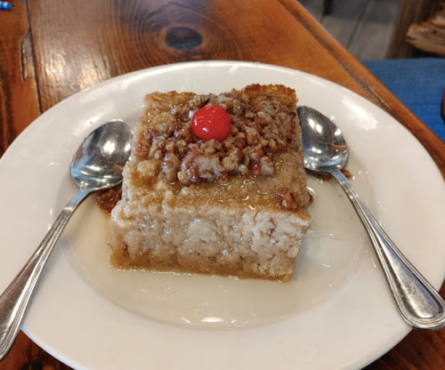 A slice of dessert topped with nuts and a cherry, served on a white plate with two spoons on a wooden table.