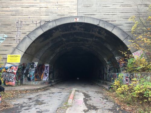 A dark tunnel entrance with graffiti-covered walls and autumn foliage nearby. A caution sign is visible.
